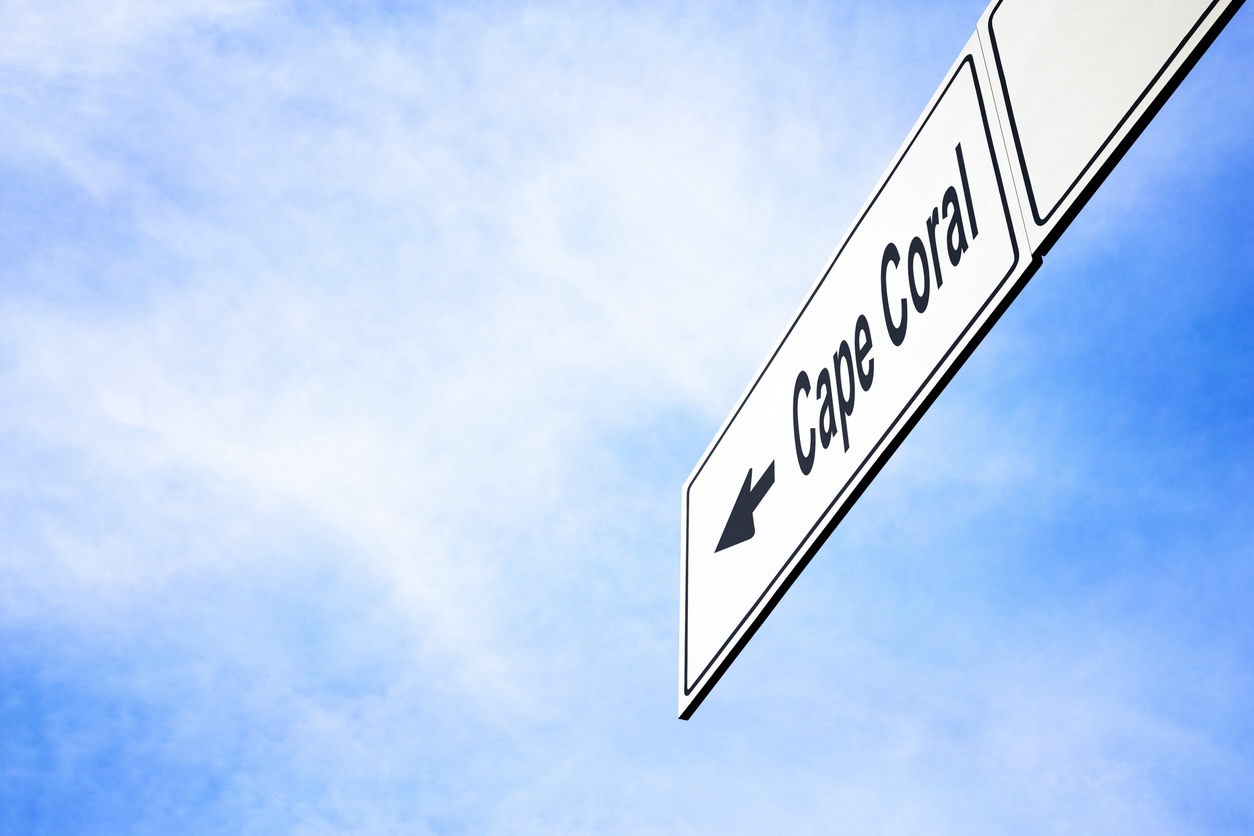 White signboard with an arrow pointing left towards Cape Coral, Florida, USA, against a hazy blue sky in a concept of travel, navigation and direction. Path included for the signboard