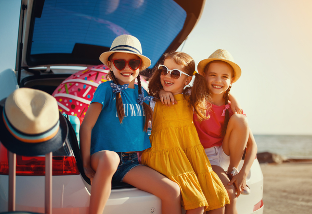Three young brightly dressed girls on a road trip with beach gear and suitcases in car trunk