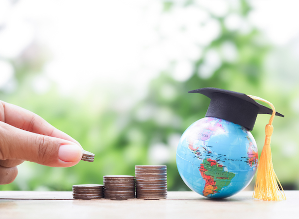 a small blue world globe with a student graduate black hat and a hand piling up coins
